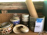 Photo showing assorted small garden pots including a mosaic ceramic pot, floral small pot, gray pencil holder pot, stacked white pots, and large conch shell. Positioned on a wooden shelf with pegboard background.