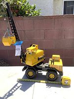 Large yellow Tonka crane truck outdoors resting on a flat surface with measuring tape showing approximately 28.8 inches in length. Black perforated crane boom equipped with yellow bucket hanging from cables.