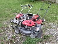 Three used gasoline-powered push lawn mowers placed on grass and gravel surface, showing front and side views with visible wear on blades, frames, and engine covers.