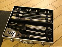 Open case showing barbecue tools neatly secured inside with black elastic straps on the lid and base including spatula, fork, tongs, and skewers.