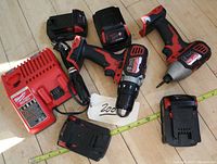 Overhead view of two cordless power tools, multiple battery packs and charger laid out on wooden floor with measuring tape
