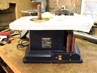 Front view of the oscillating spindle sander on a wooden workbench showing the sanding spindle attached to the table and three additional sanding spindles stored on the side.