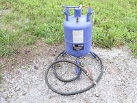 Full view of blue cylindrical 5 gallon pressure sandblaster with hose coiled around base on gravel and grass background.