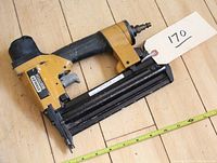 Multiple angles of the yellow and black Stanley Bostitch BT50B brad nailer sitting on a wood floor with measuring tape for scale. The tool includes safety labels and an air hose connection at rear.