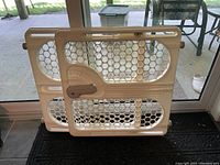 Plastic baby safety gate with a locking handle and hexagonal pattern, photographed in front of glass door.