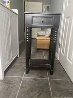 Front view of the black wooden side table showing turned legs, single drawer, and lower shelf.