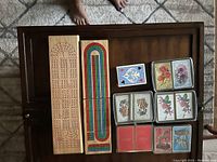 Two wooden cribbage boards and multiple decks of playing cards displayed on a dark wooden surface with a light floor background.