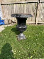 Front side view of black cast iron plant holder with detailed rim and pedestal base, sitting on green grass next to wooden fence and small blue chair.