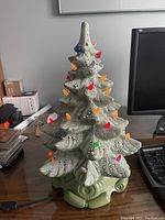 Ceramic Christmas tree with textured green branches and multicolored bulbs lit on a wooden desk beside a keyboard and monitor.