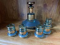 Aqua-tinted glass decanter with gold trim and stopper, plus four matching small glasses with gold accents, arranged on wooden shelf.