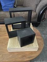 Two sets of three black floating shelves arranged on a wooden surface. Each shelf is hollow-centered square or rectangular shape with a matte black finish.