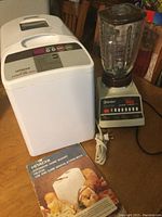 Osterizer blender with glass pitcher and control buttons beside white Hitachi bread maker with manual and cookbook on wooden table