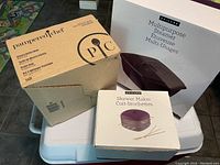 Three boxed kitchen items: Pampered Chef Batter Bowl, Epicure Multipurpose Steamer, and Epicure Skewer Maker on white cooler, angled view.