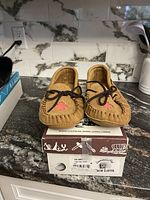 Front view of the tan suede moccasin slippers on original box showing red maple leaf detail and lace bows.