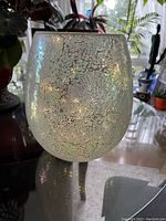 Front view of the light blue iridescent crackle glass hurricane lamp on a table in a well-lit room with plants in the background.