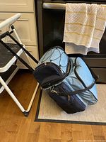 Photo of closed light blue and black soft-sided picnic carrying case with shoulder strap, placed on floor next to a chair