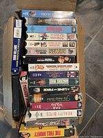 Photo of a box with approximately 40 VHS movies showing various titles lined up side by side in good condition, mostly in original cases.