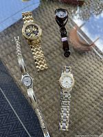 Four wristwatches arranged on glass table; gold-tone, silver-tone, two-tone, and black designs visible.