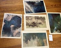 Five unframed watercolor prints laid out on wooden floor displaying abstract landscape scenes with dark and muted colors.