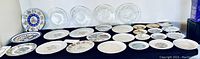 Wide view showing over 25 assorted decorative plates arranged on a black cloth, including clear glass etched plates and various floral design plates.
