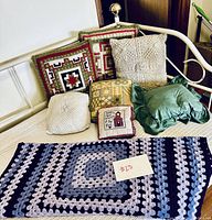 Full display of seven pillows on a white bench with a crocheted granny square blue/grey blanket placed in front with a $23 price tag