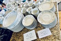 Multiple pieces of Diane Fine Porcelain China including plates, cups, saucers, and large serving bowls arranged on a countertop. Each piece has a beige and gray floral and scroll pattern with a white base.