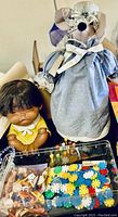 Two dolls displayed along with a clear box of assorted toy pieces and miniature cars