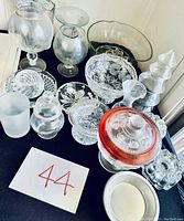 Photo of glass collection including clear pitcher, Princess House bowl, candle holder, red container, and other small glass containers arranged on black surface