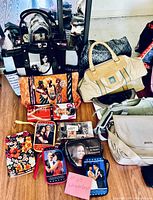 Photo showing assorted handbags, purses, and clutches arranged on floor in front of a wall, multiple styles and sizes visible, some with celebrity image prints.