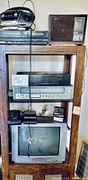 Wooden mid-century stand with various vintage electronics arranged on shelves including AM/FM receiver, TV, cassette players, and CD player.