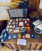 Photo of various small items arranged on wooden radio cabinet including carved magnets, film camera, battery chargers, wooden ashtray, and tray