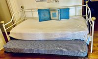 Side view of white metal framed twin trundle bed showing brass finials, top bed with mattress and cushions, and lower roll out bed mostly closed