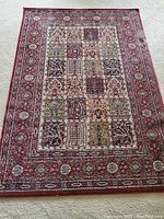 Full view of the rectangular rug showing the red border and central multi-pattern patchwork sections laid on a carpet floor.