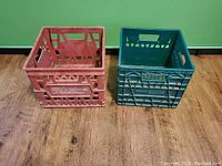 Photo showing two milk crates, one red vintage Dominion Dairies Ltd. crate and one green crate, on a wooden floor against a green wall.