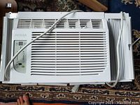 Top view of the Noma window air conditioner unit showing the front grille, control panel, power cord, and accordion side panels on a patterned carpet.