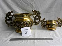 Two brass Chinese-themed decorative bowls with dragon handles side by side, ruler for scale showing measurements; larger and smaller bowls with detailed carvings and red eyes in dragon handles