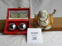 Photo showing pair of shiny metal chiming healing balls in red fabric-lined box with Chinese designs, next to ceramic Buddha figurine.