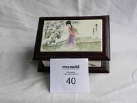 Photo showing the top of the box with ceramic panel depicting a traditional Chinese woman and bamboo trees, placed on white fabric background, with MaxSold label marked 40.