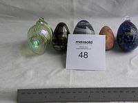 Front view of five decorative eggs with ruler for scale, showing different colors, materials and loop tops on two eggs.