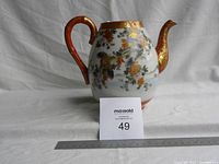 Front view of vintage porcelain teapot with floral and bird design, showing handle and spout with gold accents.