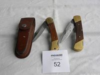 Two folding pocket knives and one brown leather sheath laid flat on a white surface, knives partially open and closed to display blades and handles.