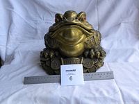 Front view of the large brass lucky frog with red eyes and coin base, ruler for size reference.