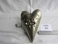 Metal heart decoration resting on white cloth, with ruler showing approximate size and a lot number card visible next to it.