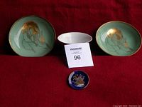 Two small Oriental ceramic dishes with gold crane bird designs on green background, displayed with MaxSold lot number card, on red cloth.