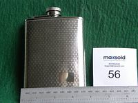Hip flask lying on a green fabric surface showing the front side with a checkered surface and a blank monogram plate. A ruler alongside shows the height and width dimensions.