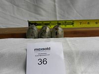 Three small decorative glass eggs displayed against a wooden backdrop, with a measuring tape showing approximately 2 inches in height for scale. A MaxSold lot number card is visible.