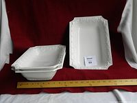 Four white rectangular ceramic serving dishes with ornate embossed borders and handles on both ends, arranged with one standing upright and the others stacked. Wooden ruler in front for scale.