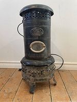 Full view of the antique black pot belly electric heating stove showing its cylindrical shape, base, perforated ventilation, and handle door.