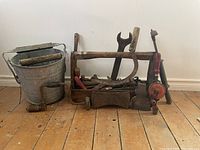 Photo showing a bucket with wooden handle, a hand brace drill, large wrench, hand saw, axe, and wooden toolbox with various tools.