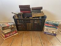 Photo showing a plastic basket filled with assorted books and two stacks of books outside it on a wooden floor